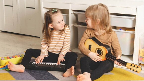 Duas crianças sentadas em um tapete no chão. A criança da esquerda está com as mãos sobre um teclado de brinquedo, enquanto a criança da direita está segurando um violão de brinquedo.
