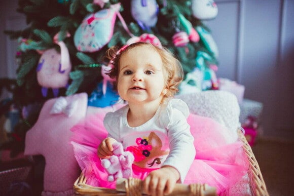 Bebê menina sorrindo enquanto brinca em um cesto perto de uma árvore de Natal decorada.